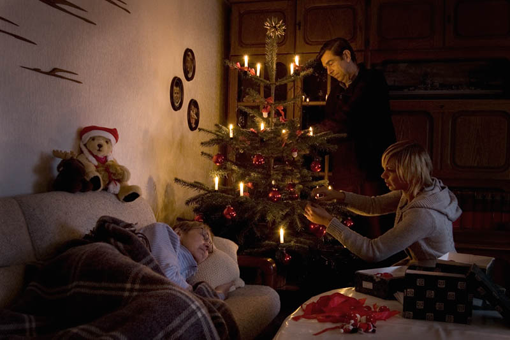 Familienszene vor einem Weihnachtsbaum, eine ältere Frau liegt zugedeckt auf dem Sofa.