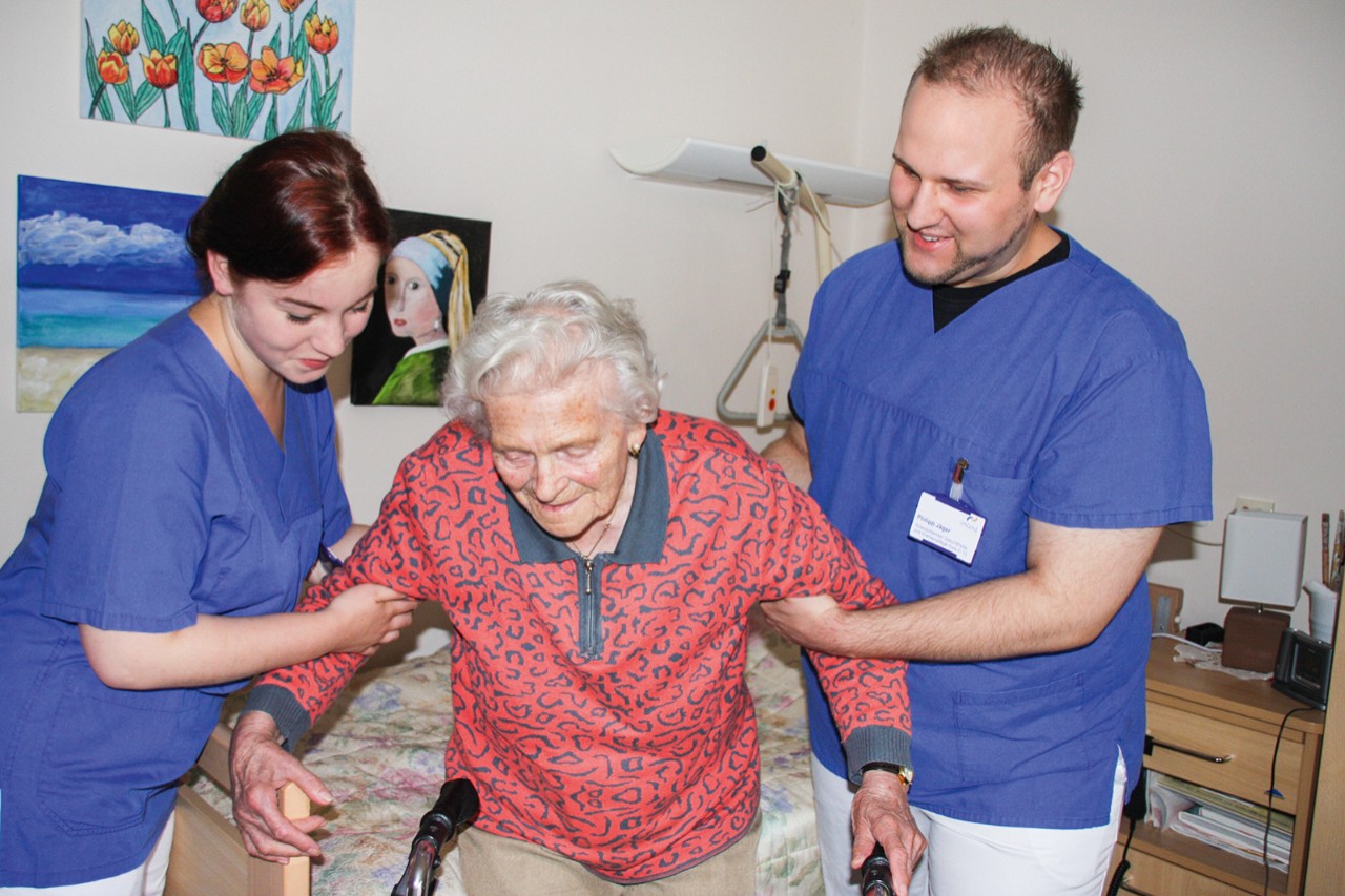 Pflegerin und Pfleger helfen alter Frau aus dem Bett.